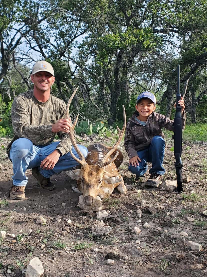 Two hunters with an Axis buck beneath trees