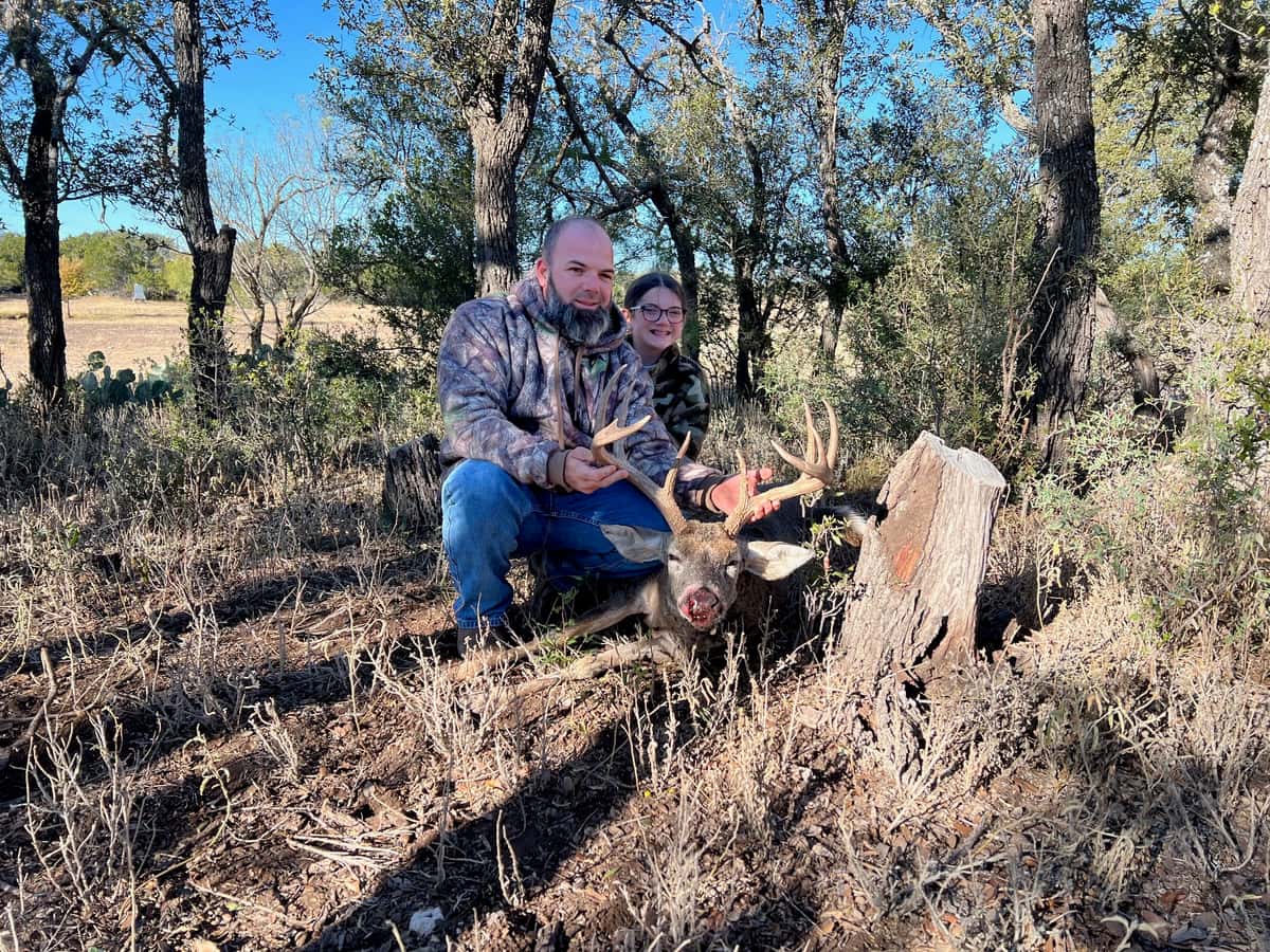 Two hunters with a whitetail buck among trees and dry grass