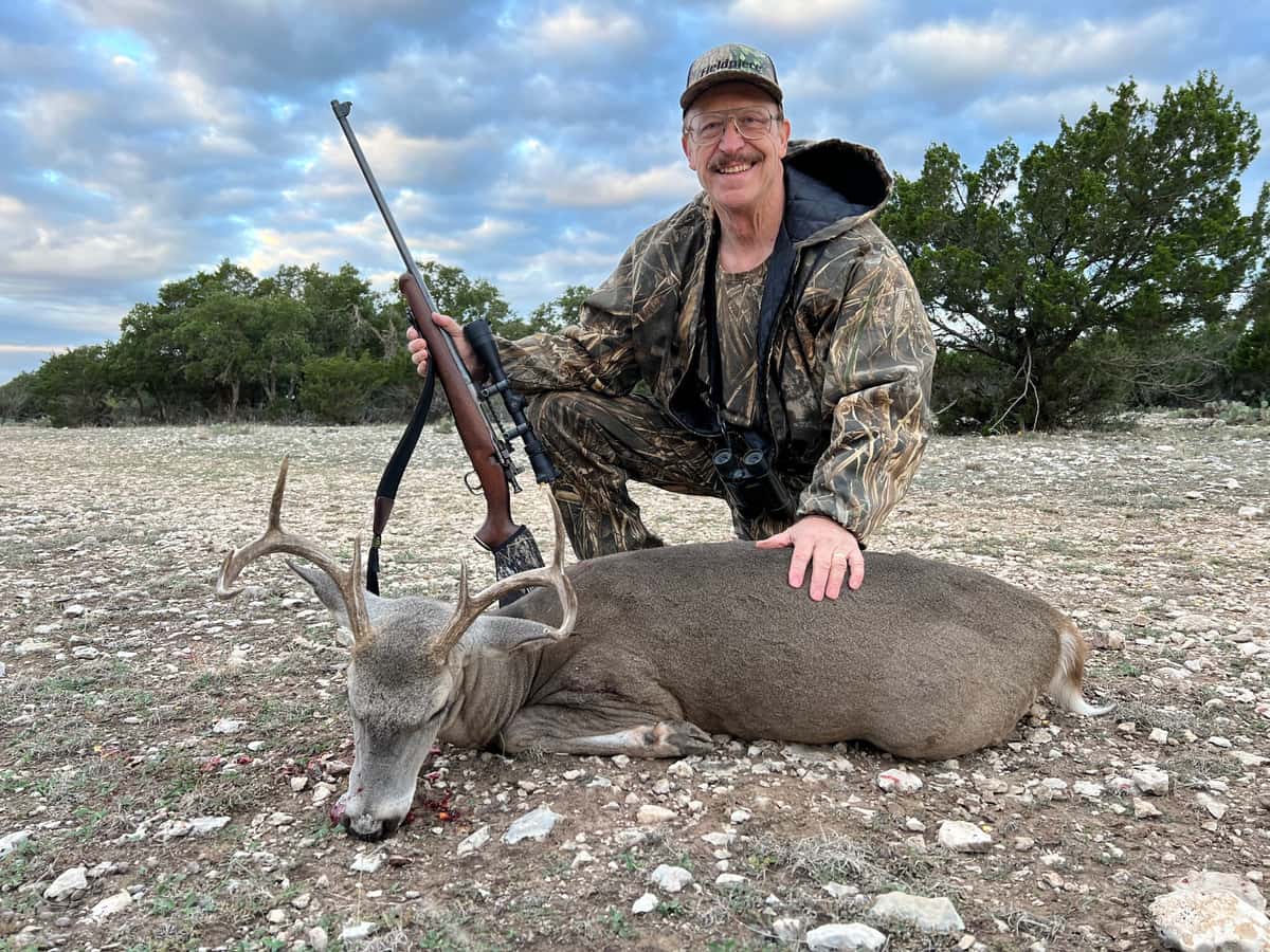 Hunter holding a rifle behind a whitetail buck on rocky ground
