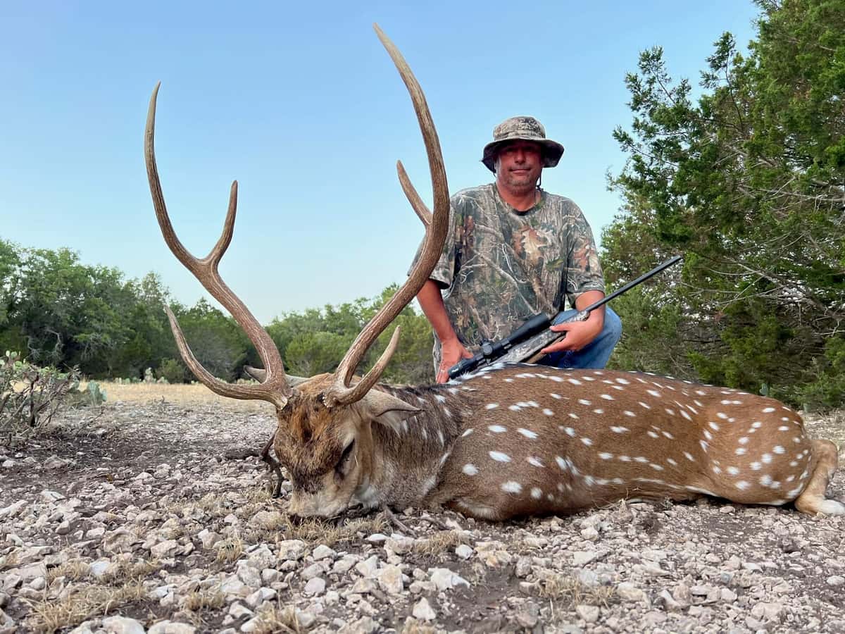 Hunter with an Axis buck on rocky ground beneath a blue sky