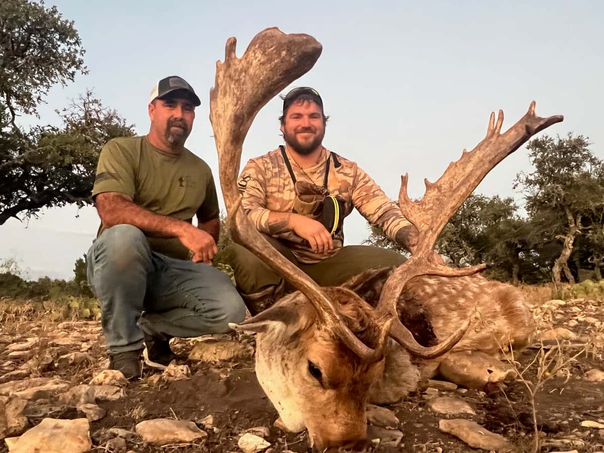 Two hunters kneeling with a fallow buck on rocky ground