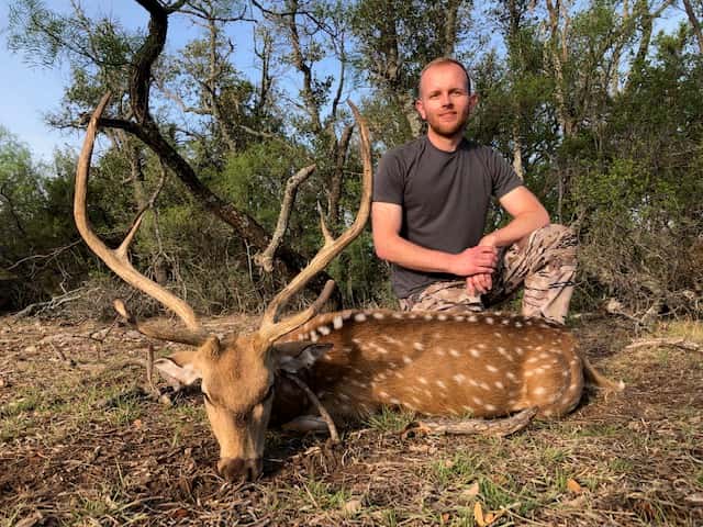 Hunter with an Axis buck in dry grass beside brush