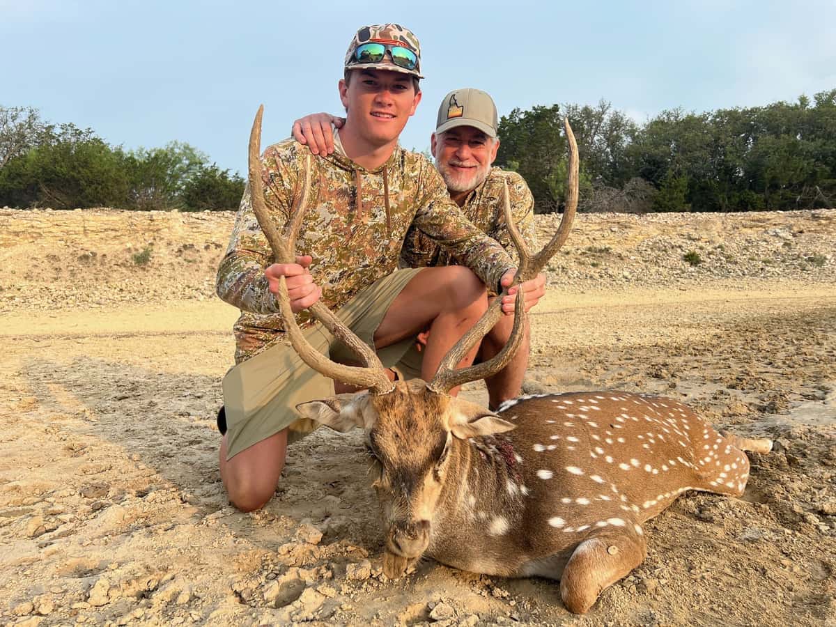 Two hunters with an Axis buck on bare ground