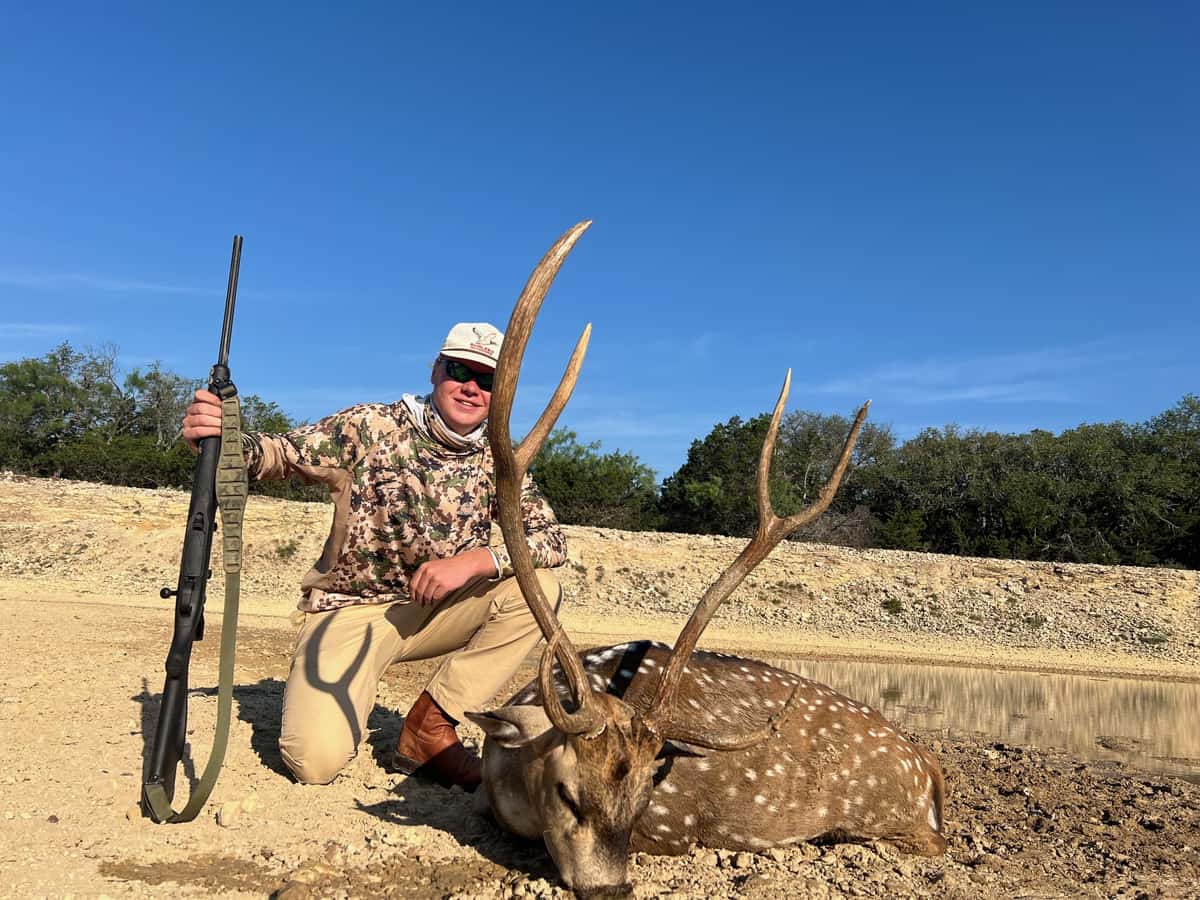 Hunter with an Axis buck and rifle on bare ground