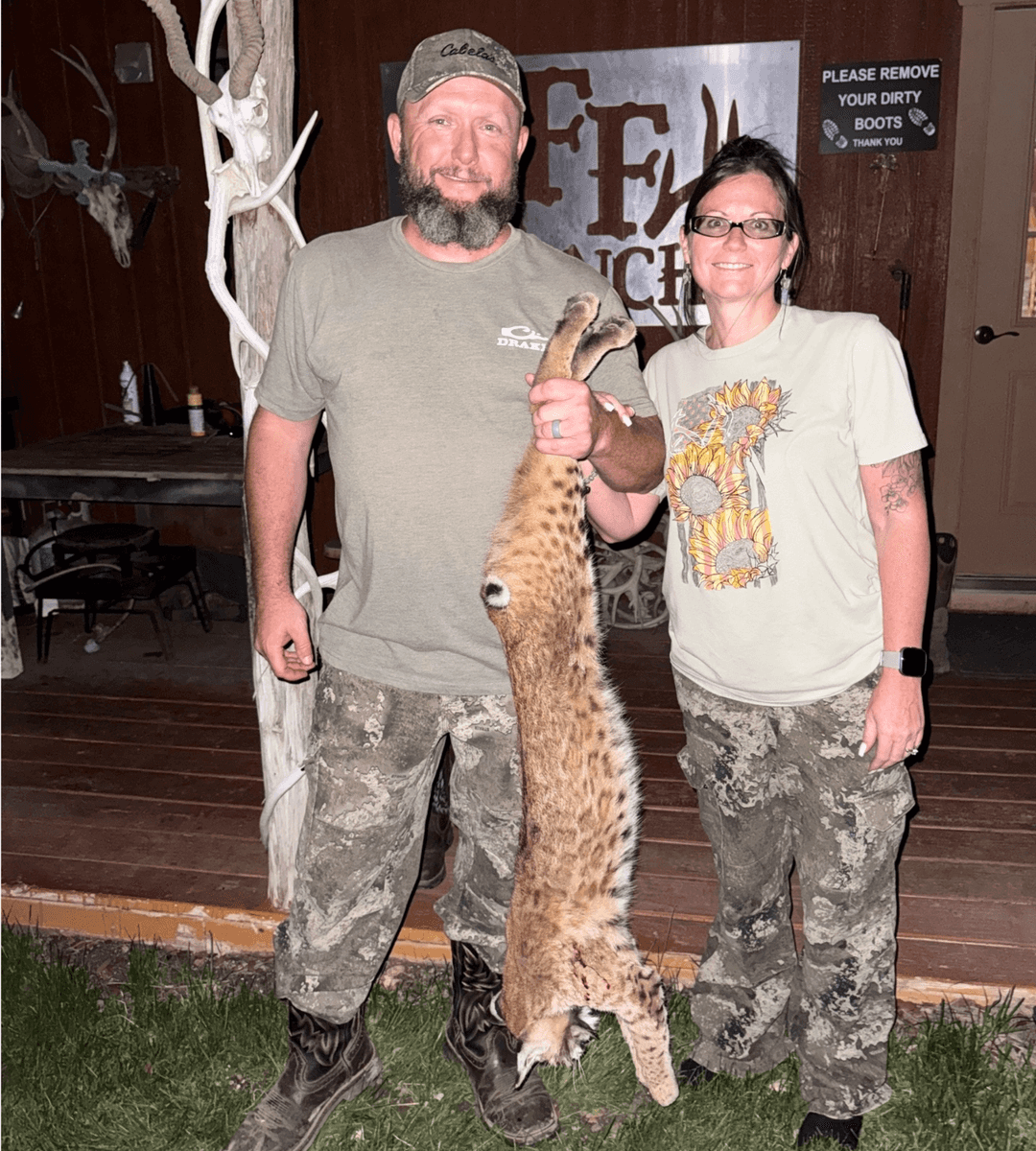 Two people holding a bobcat in front of the ranch building