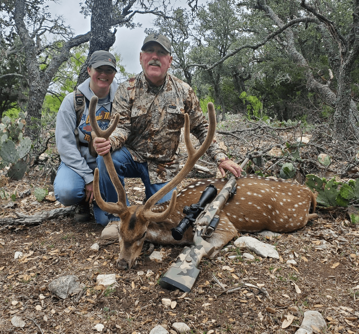 Two hunters with an Axis buck beneath trees