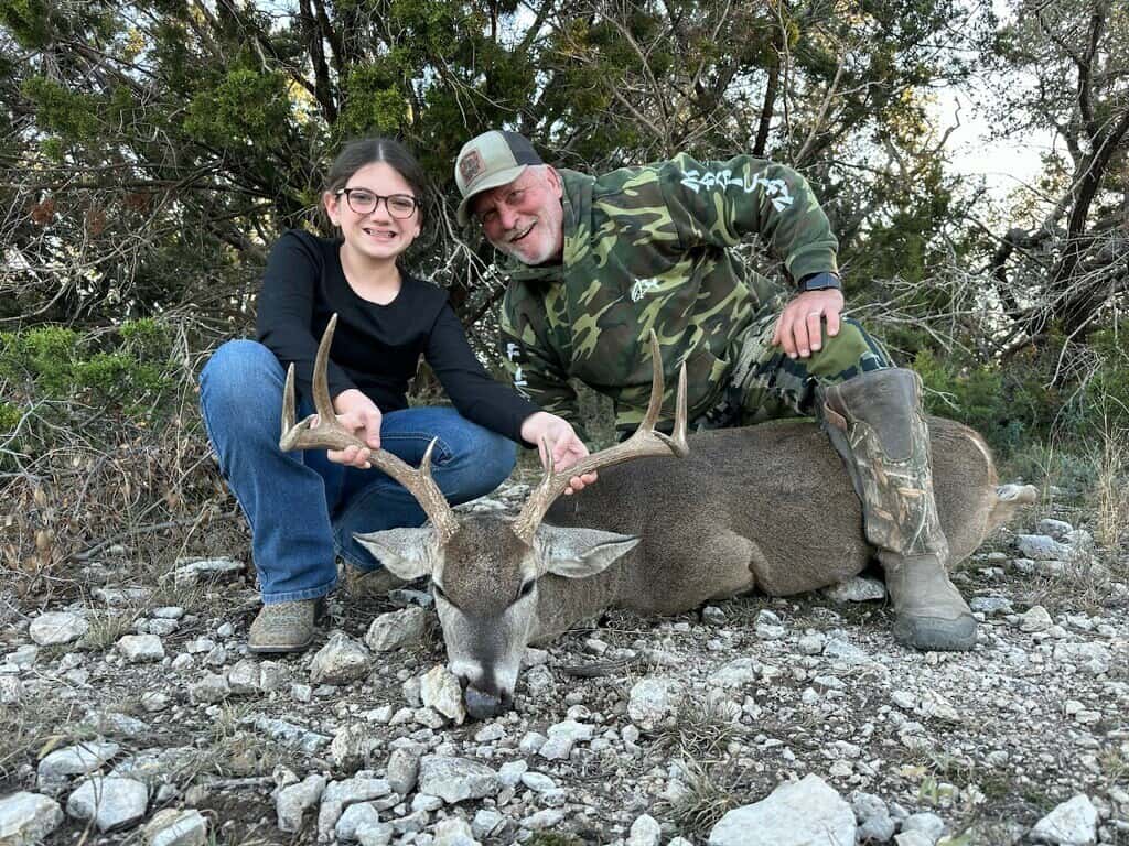 Two hunters kneeling with a whitetail buck beneath trees