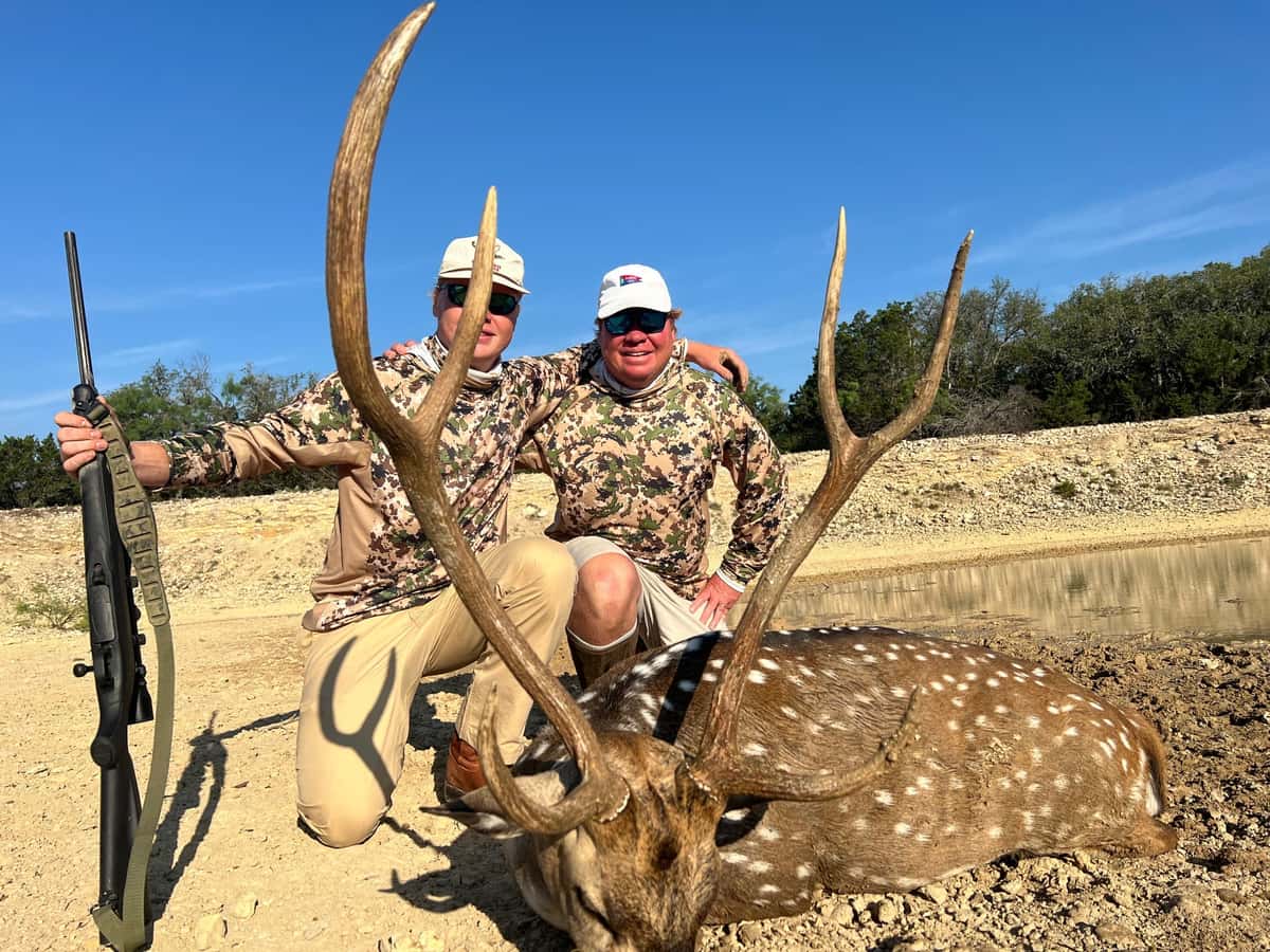 Two hunters with an Axis buck and rifle beneath a blue sky