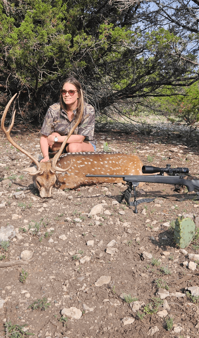 Hunter with an Axis buck and rifle beside a tree