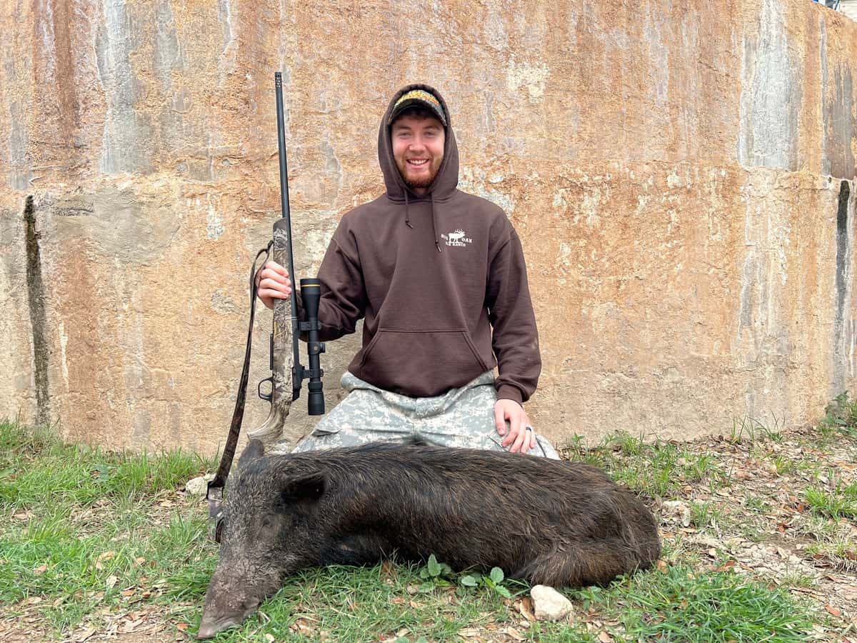 Hunter with a hog and rifle beside a round tank