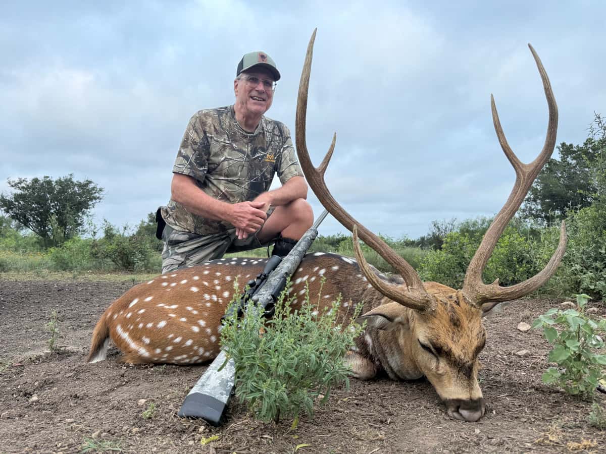 Hunter with an Axis buck and rifle on rocky ground
