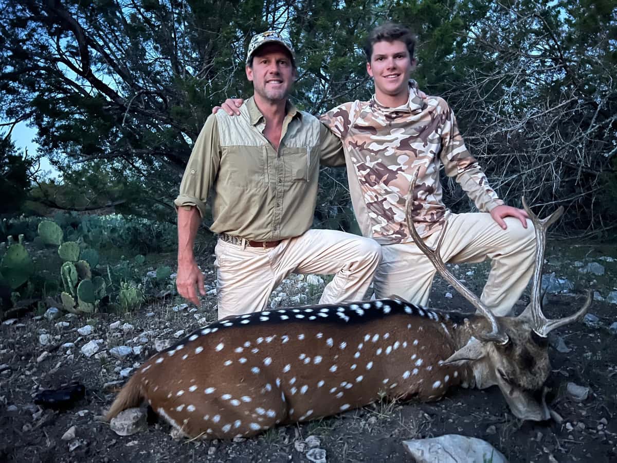 Two hunters with an Axis buck beside trees