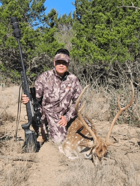 Hunter holding a rifle beside an Axis buck and cedar trees